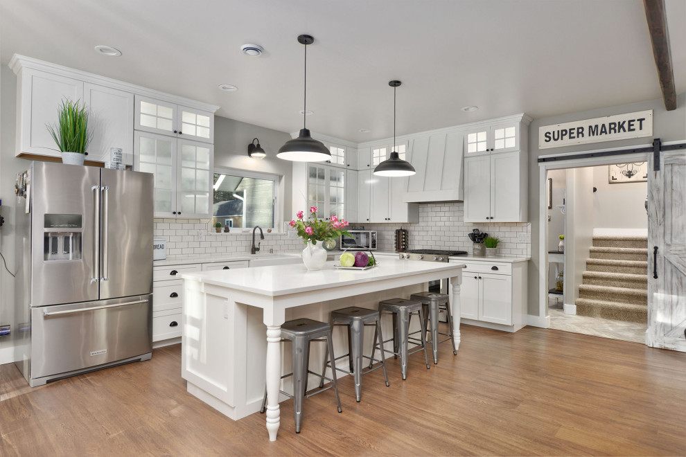 Inspiration for a country l-shaped open plan kitchen in Other with a farmhouse sink, shaker cabinets, white cabinets, white splashback, subway tile splashback, stainless steel appliances, medium hardwood floors, with island, brown floor, white benchtop and exposed beam.