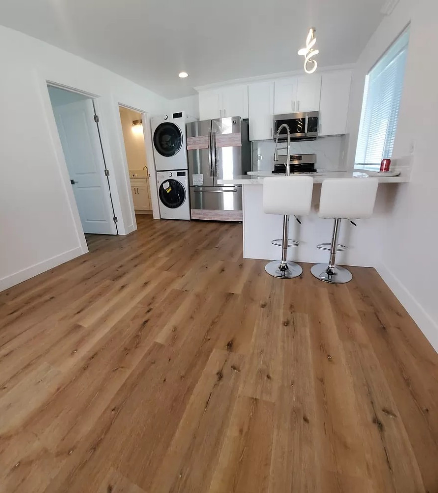 Small contemporary l-shaped separate kitchen in Los Angeles with a farmhouse sink, shaker cabinets, white cabinets, white splashback, stainless steel appliances, vinyl floors, a peninsula, brown floor and white benchtop.