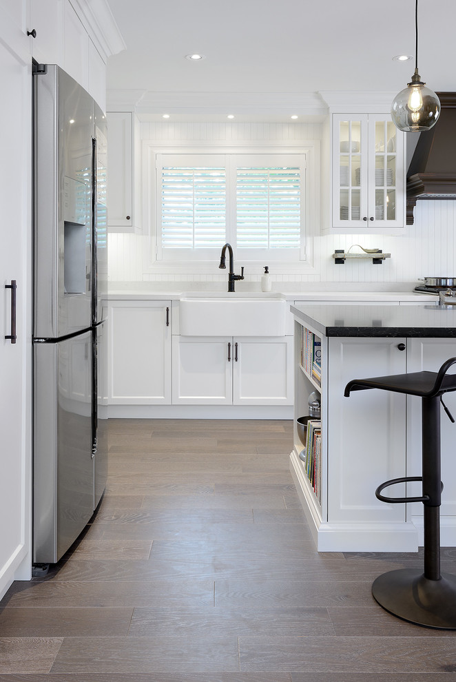 Photo of a mid-sized country l-shaped eat-in kitchen in Toronto with a farmhouse sink, white cabinets, quartz benchtops, white splashback, stainless steel appliances, medium hardwood floors and with island.