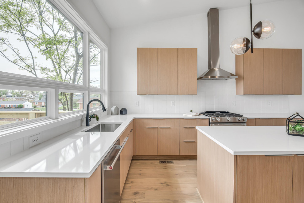 Design ideas for a mid-sized midcentury l-shaped open plan kitchen in Nashville with an undermount sink, flat-panel cabinets, light wood cabinets, quartz benchtops, white splashback, porcelain splashback, stainless steel appliances, light hardwood floors, with island and white benchtop.