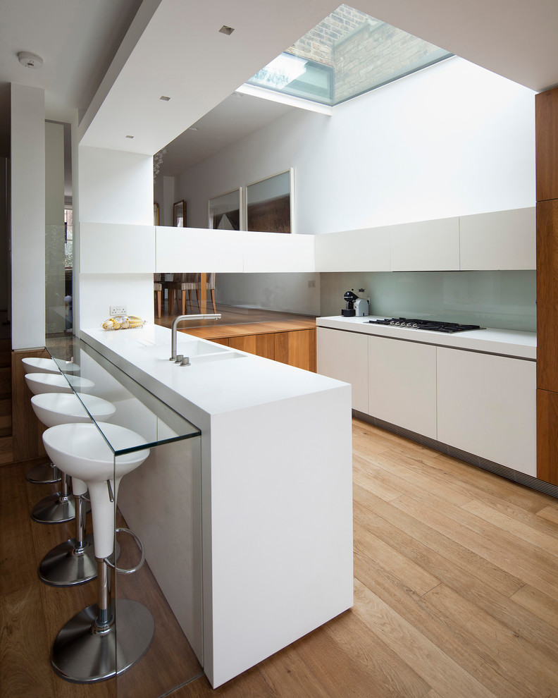 Inspiration for a contemporary galley kitchen in London with a double-bowl sink, flat-panel cabinets, white cabinets, grey splashback, glass sheet splashback, light hardwood floors, a peninsula and white benchtop.