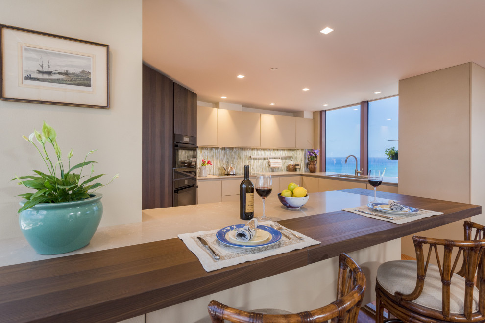 This is an example of a small modern u-shaped eat-in kitchen in Hawaii with an undermount sink, flat-panel cabinets, beige cabinets, wood benchtops, multi-coloured splashback, glass tile splashback, black appliances, medium hardwood floors, a peninsula, brown floor and brown benchtop.