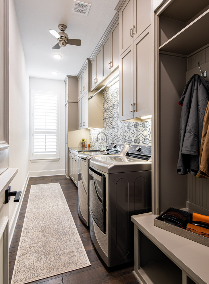 Photo of a mid-sized transitional single-wall utility room in Dallas with a farmhouse sink, shaker cabinets, grey cabinets, white walls, medium hardwood floors, a side-by-side washer and dryer, brown floor and black benchtop.
