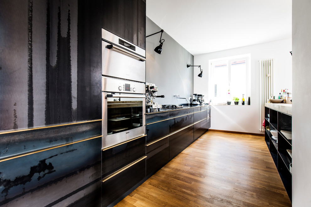 Photo of a mid-sized contemporary galley open plan kitchen in Munich with flat-panel cabinets, black cabinets, medium hardwood floors, wood benchtops, grey splashback, stainless steel appliances, an integrated sink, no island and brown floor.