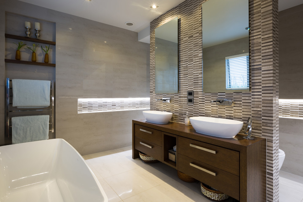 Design ideas for a large contemporary master bathroom in Auckland with medium wood cabinets, a freestanding tub, a one-piece toilet, multi-coloured tile, multi-coloured walls, porcelain floors, a vessel sink, wood benchtops, mosaic tile, beige floor, brown benchtops and flat-panel cabinets.