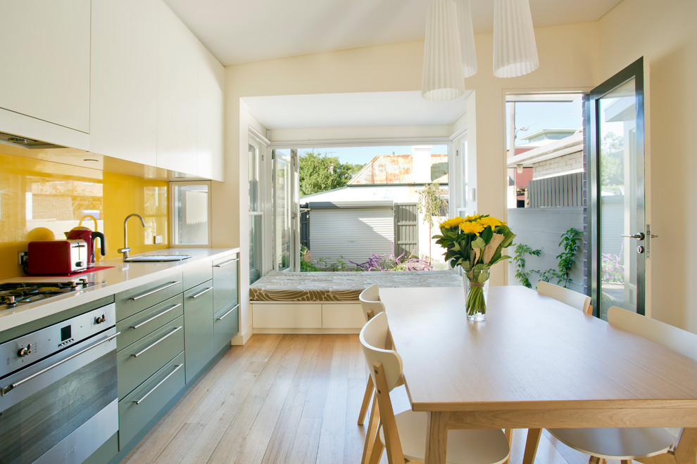This is an example of a small contemporary single-wall eat-in kitchen in Sydney with flat-panel cabinets, stainless steel appliances, yellow splashback, glass sheet splashback, green cabinets, a single-bowl sink, light hardwood floors and no island.
