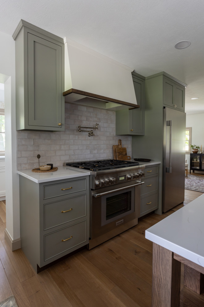 Inredning av ett vit vitt kök, med en rustik diskho, skåp i shakerstil, gröna skåp, vitt stänkskydd, stänkskydd i stenkakel, rostfria vitvaror, mellanmörkt trägolv, en köksö och brunt golv