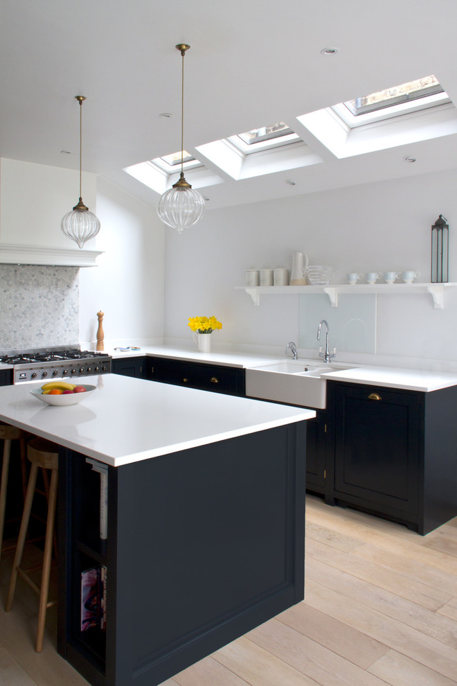 This is an example of a mid-sized transitional l-shaped kitchen in London with a farmhouse sink, recessed-panel cabinets, blue cabinets, glass sheet splashback, stainless steel appliances, light hardwood floors, with island, beige floor and white benchtop.
