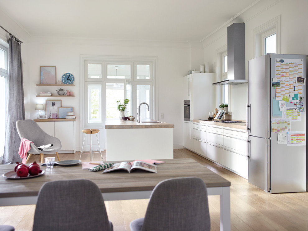 Photo of a large scandinavian galley open plan kitchen in Hamburg with a drop-in sink, white cabinets, wood benchtops, white splashback, glass sheet splashback, stainless steel appliances, light hardwood floors, with island, brown floor and flat-panel cabinets.