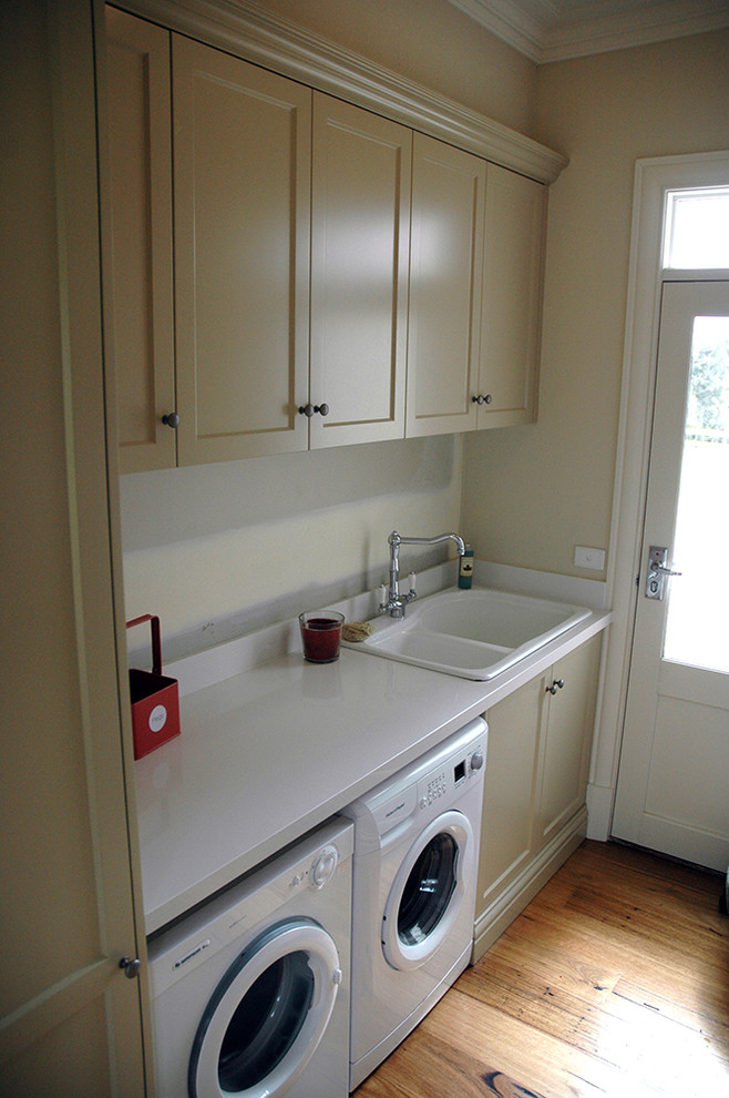 Inspiration for a traditional galley laundry room in Melbourne with a farmhouse sink, recessed-panel cabinets, beige cabinets, beige walls, medium hardwood floors and a side-by-side washer and dryer.