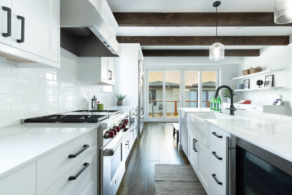 This is an example of a large arts and crafts galley open plan kitchen in Seattle with a farmhouse sink, shaker cabinets, white cabinets, quartz benchtops, white splashback, cement tile splashback, stainless steel appliances, medium hardwood floors, with island, multi-coloured floor, white benchtop and exposed beam.