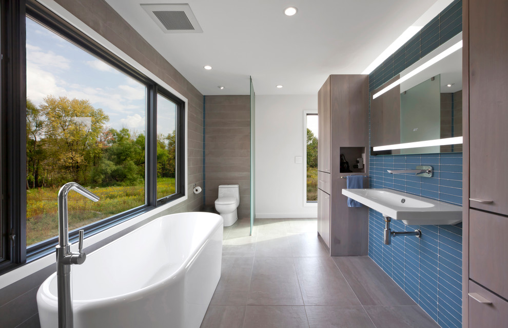 Photo of a mid-sized contemporary master bathroom in Indianapolis with flat-panel cabinets, medium wood cabinets, a freestanding tub, an open shower, a one-piece toilet, blue tile, porcelain tile, white walls, porcelain floors, a wall-mount sink, grey floor and an open shower.