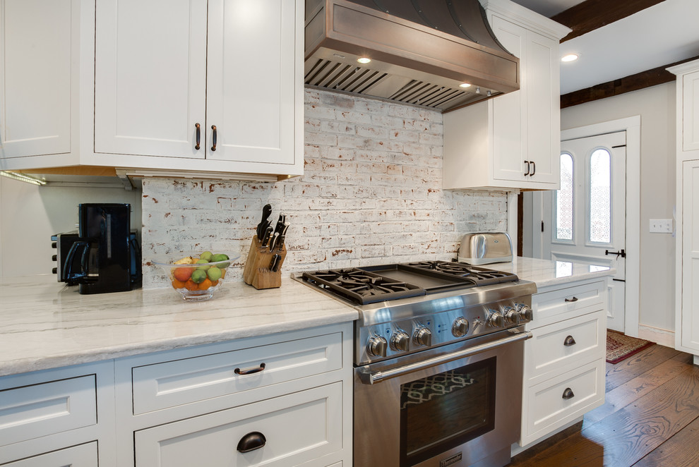 Design ideas for a mid-sized country l-shaped open plan kitchen in Providence with a farmhouse sink, beaded inset cabinets, white cabinets, quartzite benchtops, white splashback, brick splashback, stainless steel appliances, medium hardwood floors and with island.