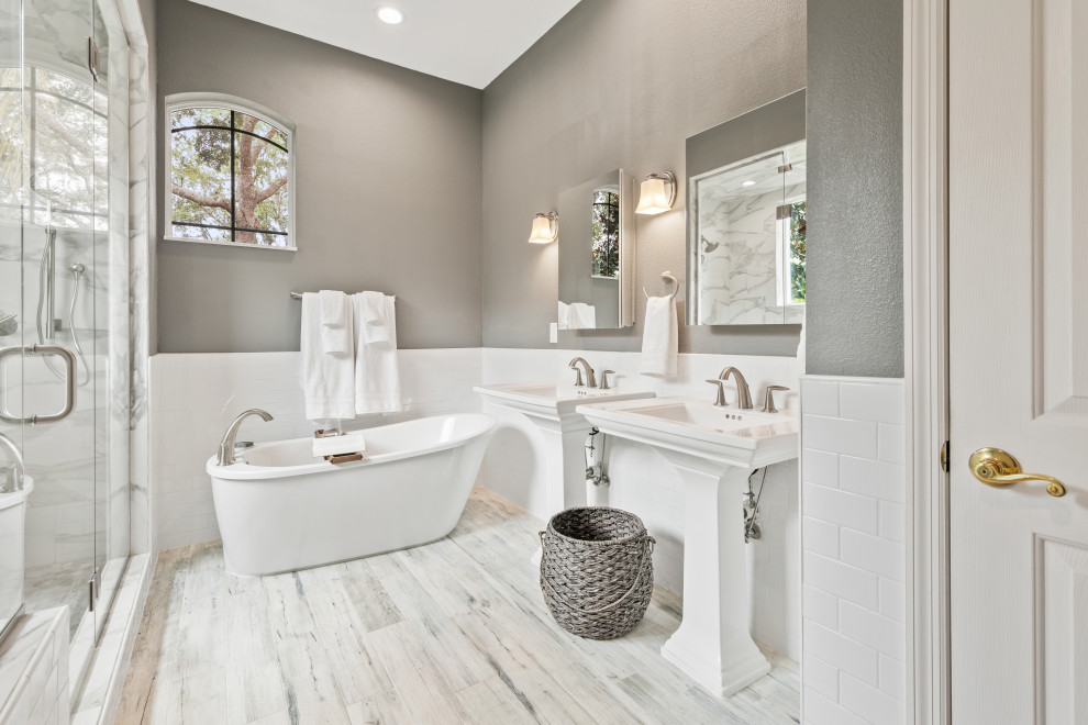 Photo of a transitional master bathroom in Orlando with a freestanding tub, a one-piece toilet, white tile, grey walls, wood-look tile, a pedestal sink, grey floor, a shower seat, a double vanity and a freestanding vanity.