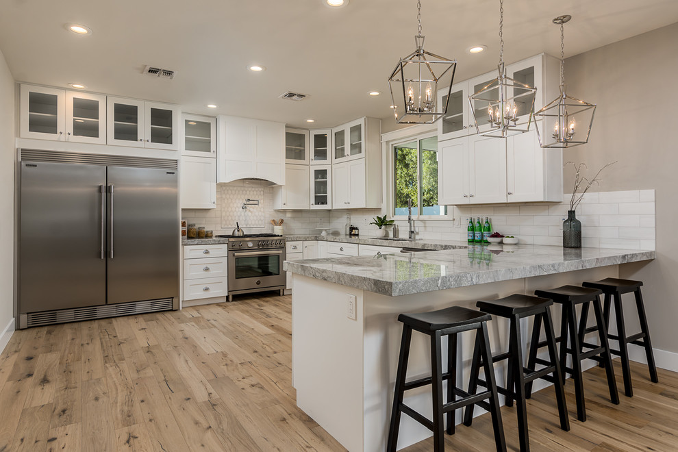 Inspiration for a transitional l-shaped kitchen in Phoenix with an undermount sink, shaker cabinets, white cabinets, white splashback, subway tile splashback, stainless steel appliances, medium hardwood floors, a peninsula, brown floor and grey benchtop.