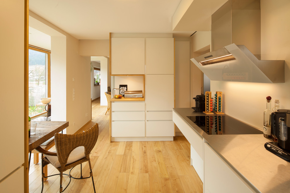 This is an example of a mid-sized contemporary single-wall eat-in kitchen in Other with white cabinets, stainless steel benchtops, medium hardwood floors, no island and flat-panel cabinets.