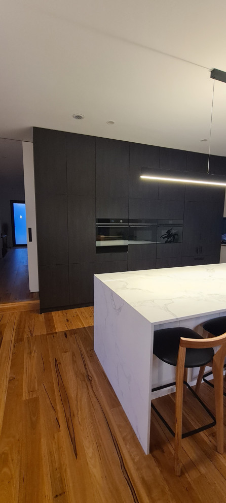 Design ideas for a large contemporary galley open plan kitchen in Geelong with a double-bowl sink, dark wood cabinets, solid surface benchtops, white splashback, ceramic splashback, black appliances, medium hardwood floors, with island and white benchtop.