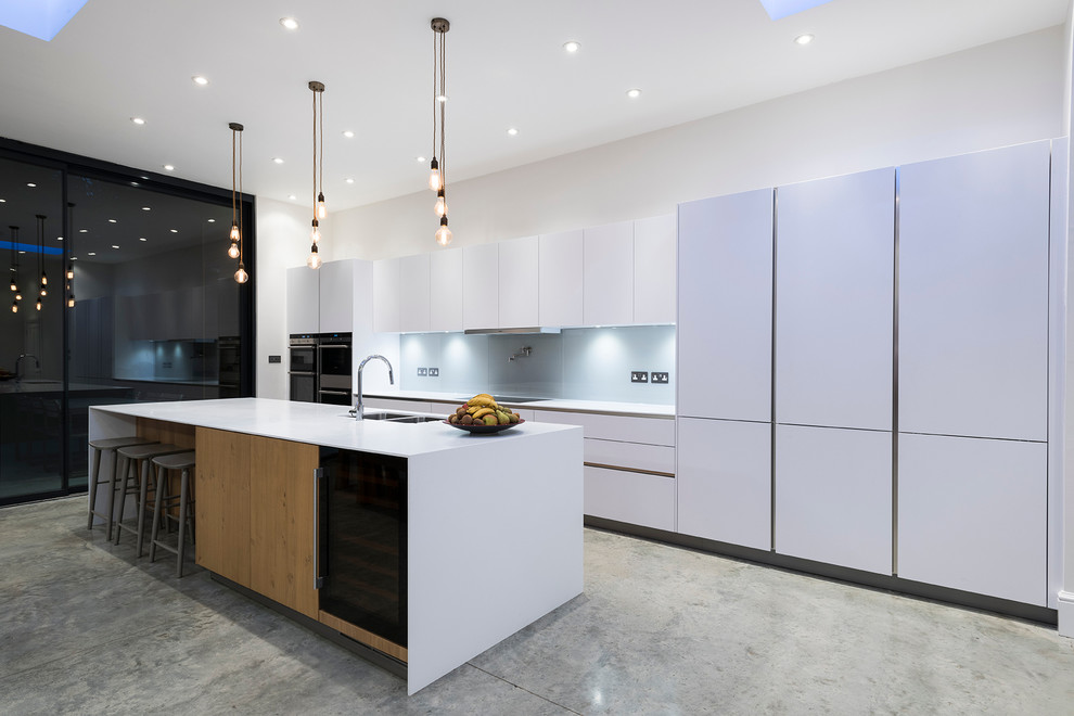 Photo of a large contemporary galley open plan kitchen in London with a drop-in sink, flat-panel cabinets, white cabinets, white splashback, with island, black appliances, concrete floors and glass sheet splashback.