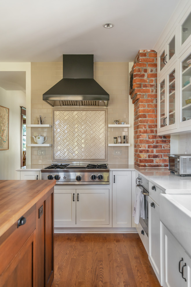 This is an example of a mid-sized arts and crafts l-shaped eat-in kitchen in Seattle with a farmhouse sink, glass-front cabinets, beige splashback, with island, white benchtop, stainless steel appliances, white cabinets, quartz benchtops, ceramic splashback and medium hardwood floors.