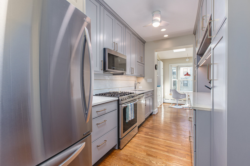 This is an example of a small contemporary galley kitchen pantry in DC Metro with a submerged sink, shaker cabinets, grey cabinets, engineered stone countertops, white splashback, glass tiled splashback, stainless steel appliances, medium hardwood flooring, brown floors and white worktops.