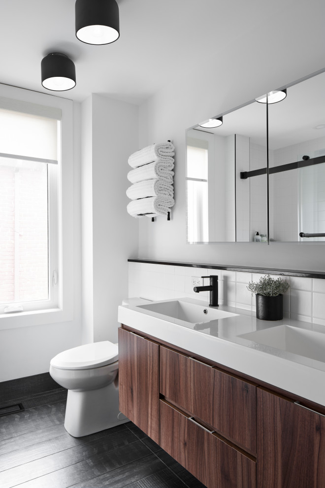 Design ideas for a mid-sized contemporary kids bathroom in Toronto with flat-panel cabinets, medium wood cabinets, an alcove tub, an alcove shower, a one-piece toilet, white tile, porcelain tile, white walls, porcelain floors, an integrated sink, solid surface benchtops, black floor, a sliding shower screen, white benchtops, a double vanity and a floating vanity.