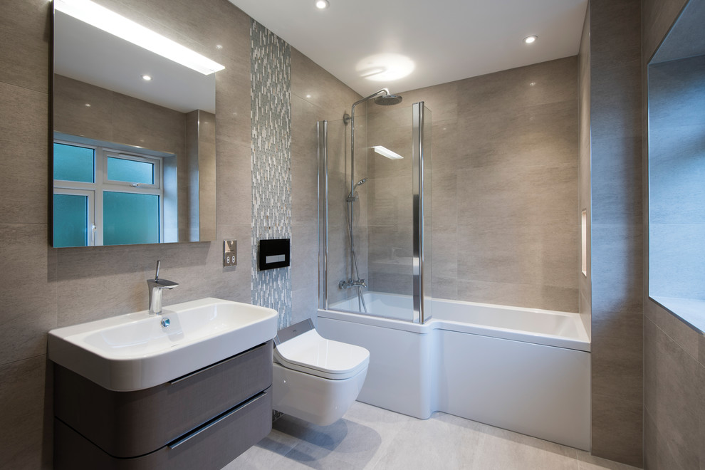Mid-sized contemporary bathroom in West Midlands with grey cabinets, a shower/bathtub combo, a wall-mount toilet, flat-panel cabinets, an alcove tub, gray tile, grey walls, a wall-mount sink and grey floor.