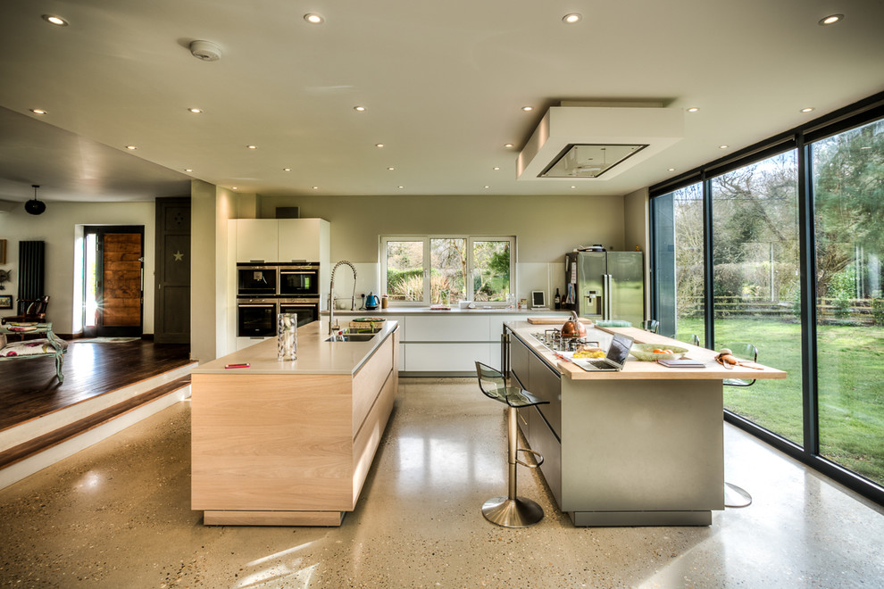 This is an example of a large contemporary u-shaped open plan kitchen in Berkshire with a double-bowl sink, flat-panel cabinets, light wood cabinets, white splashback, glass tile splashback, stainless steel appliances, concrete floors, grey floor, quartz benchtops and multiple islands.