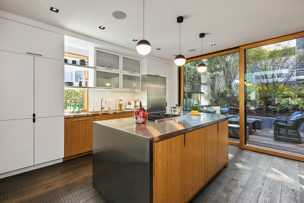 This is an example of an eclectic single-wall open plan kitchen in San Francisco with flat-panel cabinets, medium wood cabinets, stainless steel benchtops, white splashback, stone slab splashback, stainless steel appliances, dark hardwood floors, with island, brown floor, grey benchtop and an integrated sink.