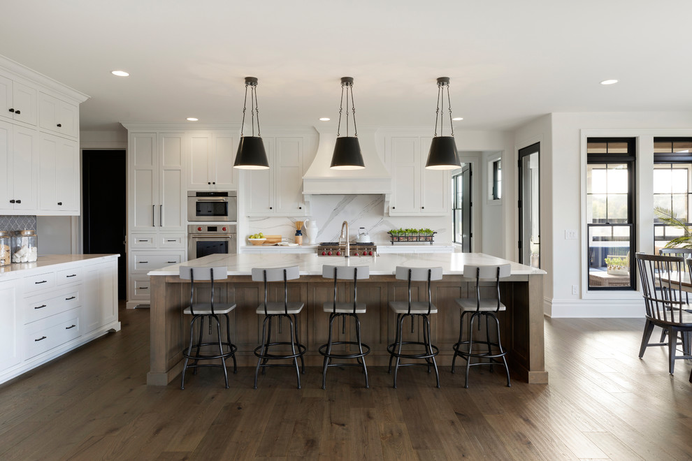 Photo of a country l-shaped eat-in kitchen in Minneapolis with recessed-panel cabinets, white cabinets, white splashback, stone slab splashback, stainless steel appliances, medium hardwood floors, with island, brown floor and white benchtop.