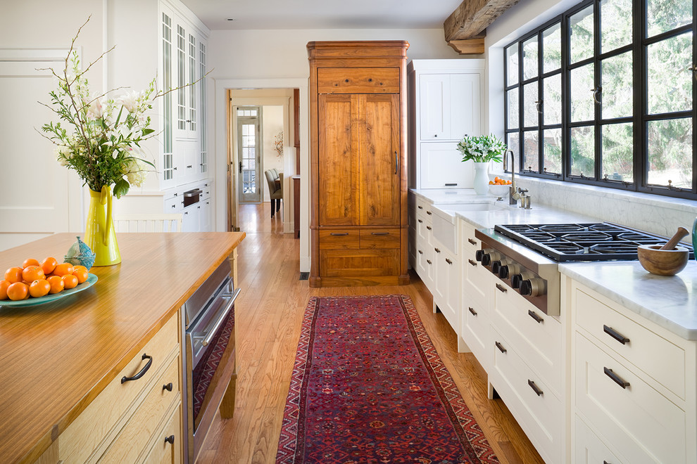 Traditional kitchen in Kansas City with glass-front cabinets, wood benchtops, panelled appliances, white cabinets, stone slab splashback and a farmhouse sink.