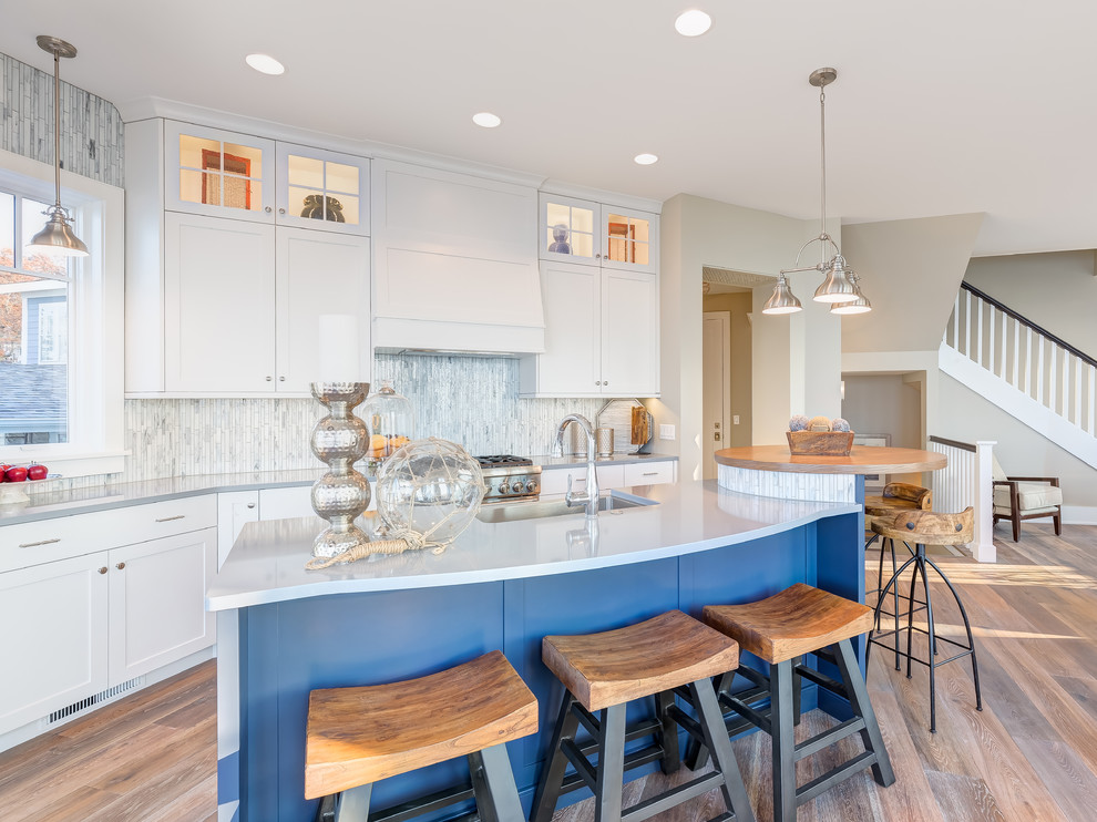 Photo of a large beach style galley open plan kitchen in Chicago with shaker cabinets, white cabinets, matchstick tile splashback, light hardwood floors, with island, a drop-in sink, quartzite benchtops, white splashback and stainless steel appliances.