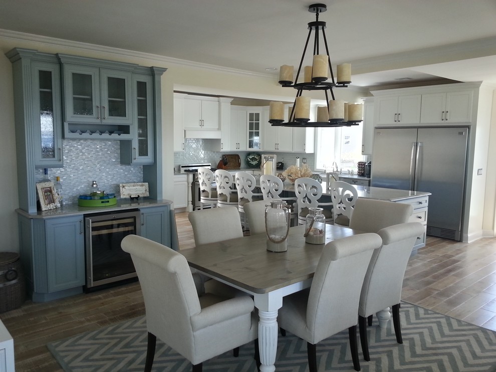 This is an example of a mid-sized beach style l-shaped eat-in kitchen in New York with shaker cabinets, white cabinets, with island, a farmhouse sink, white splashback, ceramic splashback, stainless steel appliances and light hardwood floors.