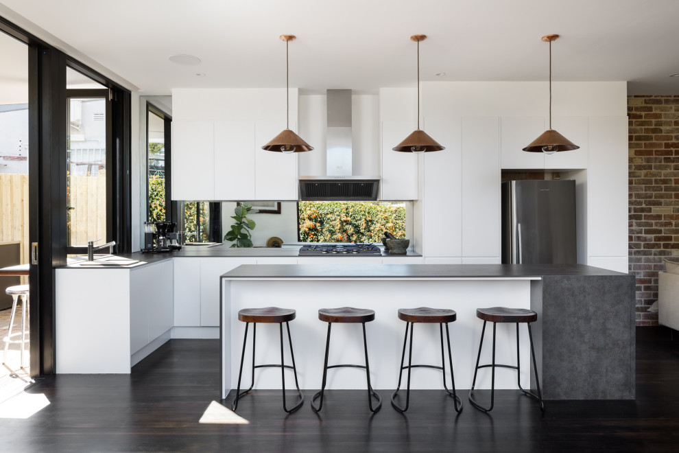 Contemporary galley open plan kitchen in Sydney with a drop-in sink, white cabinets, solid surface benchtops, mirror splashback, stainless steel appliances, painted wood floors, with island, black floor and grey benchtop.
