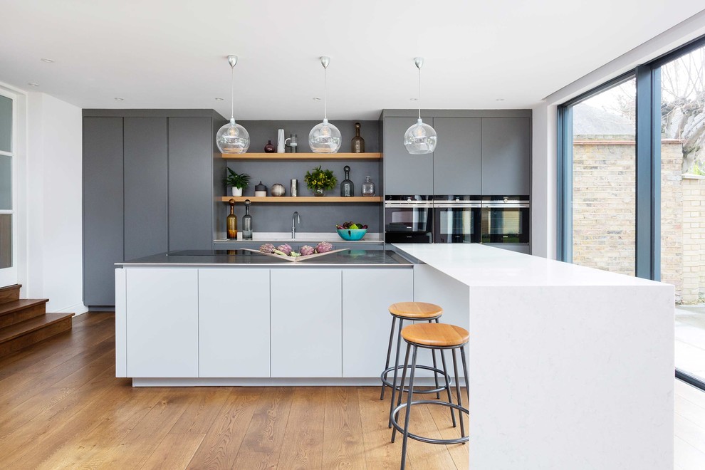 This is an example of a mid-sized contemporary single-wall kitchen in London with flat-panel cabinets, stainless steel appliances, light hardwood floors and with island.
