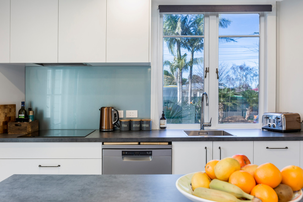 Mid-sized modern l-shaped eat-in kitchen in Auckland with a drop-in sink, flat-panel cabinets, white cabinets, laminate benchtops, glass sheet splashback, stainless steel appliances, with island, black benchtop and blue splashback.