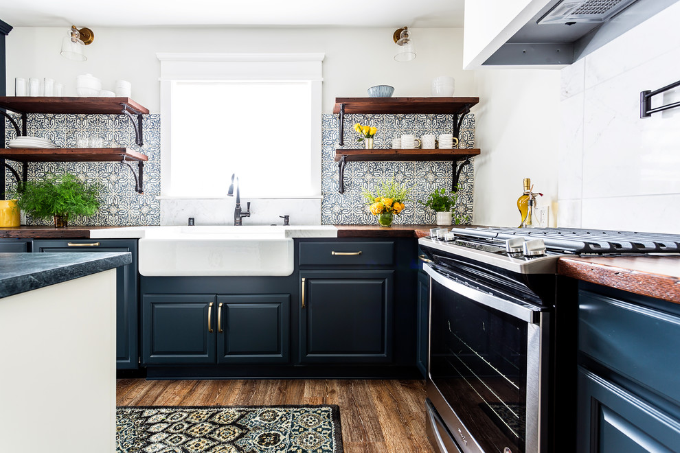 Photo of a mid-sized country l-shaped open plan kitchen in Los Angeles with a farmhouse sink, raised-panel cabinets, blue cabinets, wood benchtops, blue splashback, cement tile splashback, stainless steel appliances, laminate floors, with island, brown floor and brown benchtop.