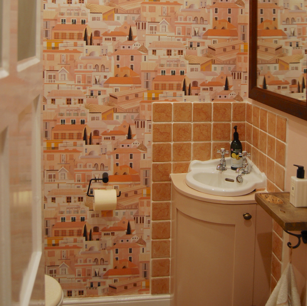 This is an example of a small country powder room in West Midlands with recessed-panel cabinets, a one-piece toilet, pink tile, ceramic tile, pink walls, ceramic floors, a wall-mount sink, wood benchtops, orange floor, pink benchtops and a built-in vanity.