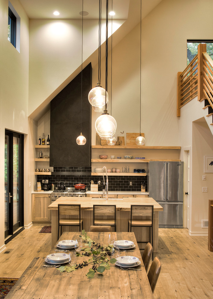 Photo of a mid-sized modern single-wall eat-in kitchen in Minneapolis with a farmhouse sink, flat-panel cabinets, light wood cabinets, solid surface benchtops, black splashback, subway tile splashback, stainless steel appliances, light hardwood floors, with island and white benchtop.