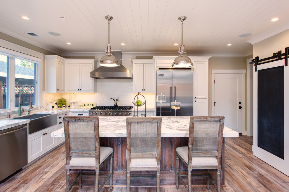 This is an example of a large country l-shaped kitchen in San Francisco with a farmhouse sink, shaker cabinets, white cabinets, marble benchtops, white splashback, stainless steel appliances, medium hardwood floors, with island and subway tile splashback.