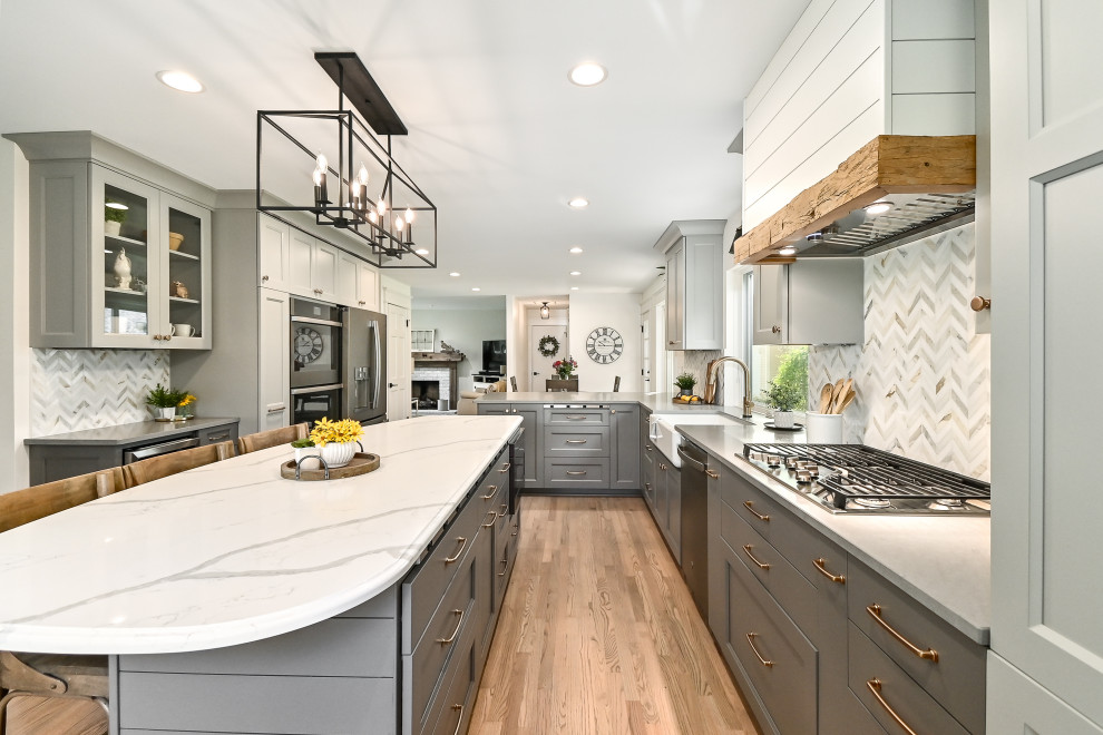 Expansive country l-shaped kitchen/diner in Milwaukee with a belfast sink, shaker cabinets, grey cabinets, engineered stone countertops, white splashback, marble splashback, stainless steel appliances, medium hardwood flooring, an island, brown floors and grey worktops.