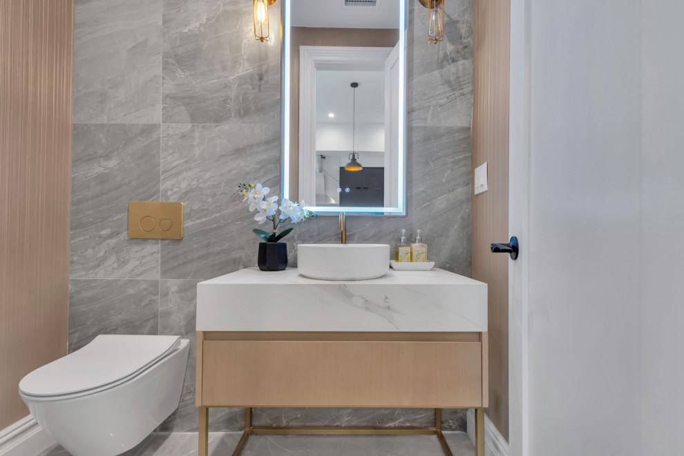 This is an example of a medium sized contemporary cloakroom in New York with flat-panel cabinets, light wood cabinets, a wall mounted toilet, grey tiles, porcelain tiles, grey walls, porcelain flooring, a pedestal sink, marble worktops, grey floors, white worktops and a freestanding vanity unit.