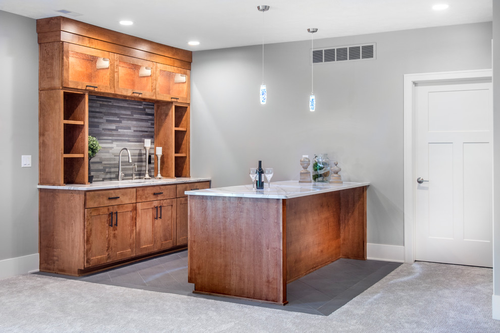 This is an example of a mid-sized arts and crafts galley seated home bar in Omaha with an undermount sink, shaker cabinets, medium wood cabinets, quartzite benchtops, grey splashback, stone tile splashback, porcelain floors, grey floor and white benchtop.