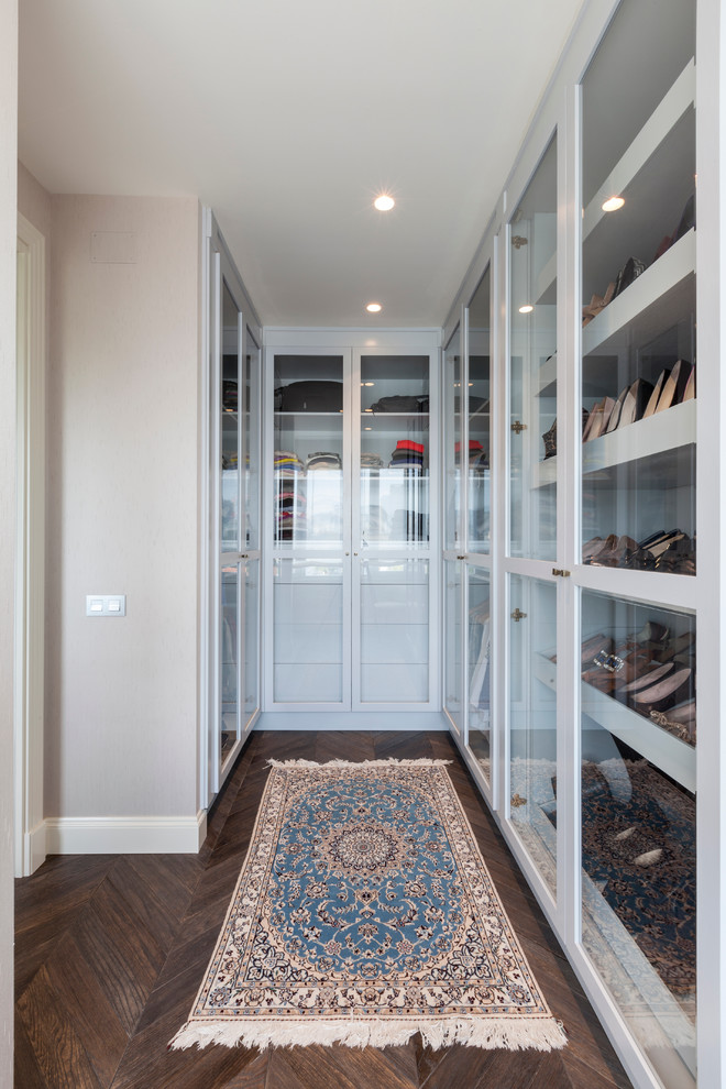 Inspiration for a mid-sized transitional gender-neutral walk-in wardrobe in London with glass-front cabinets, white cabinets and dark hardwood floors.