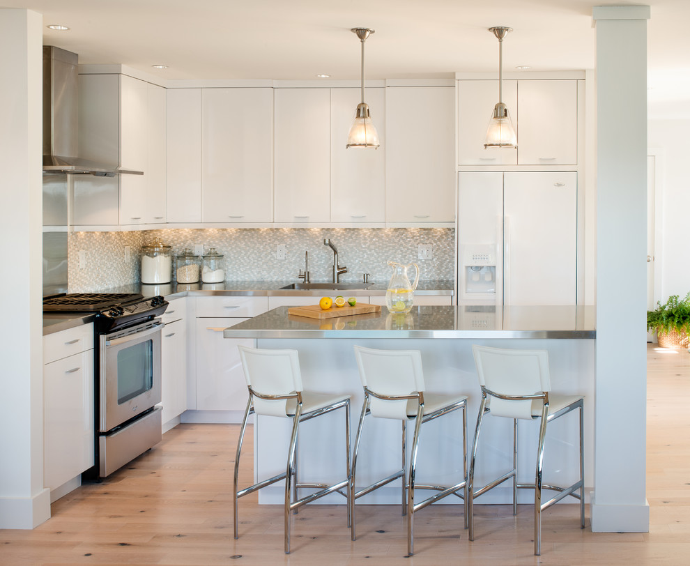 Inspiration for a beach style l-shaped kitchen in Portland Maine with an integrated sink, flat-panel cabinets, white cabinets, stainless steel benchtops, white appliances, grey splashback and mosaic tile splashback.