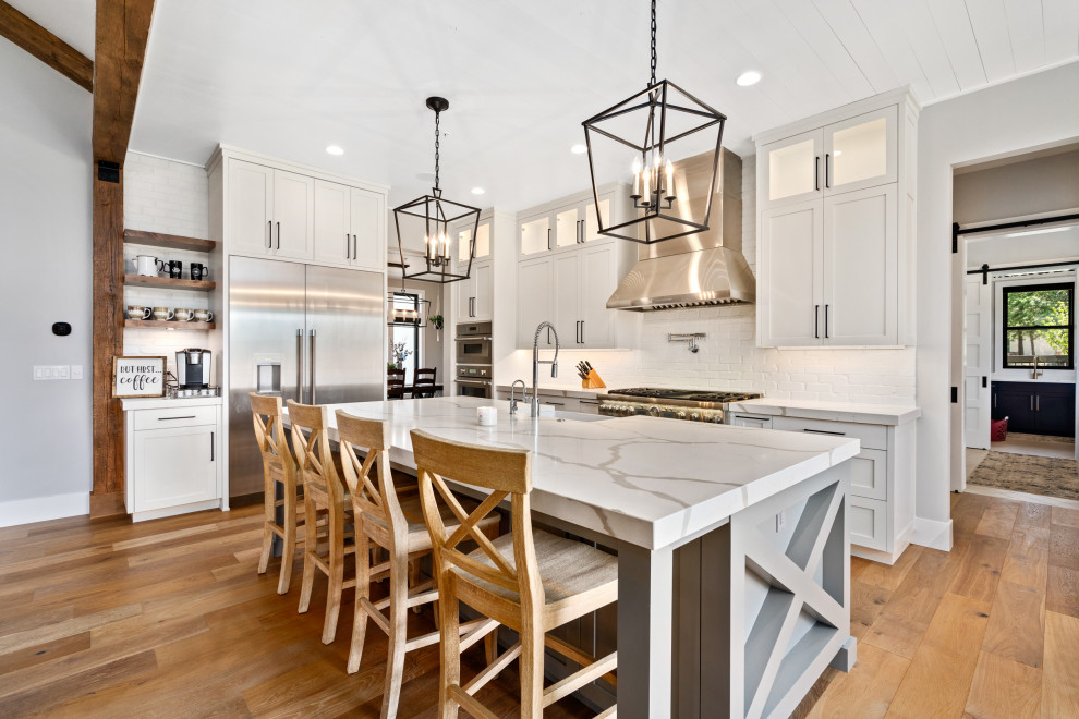 Design ideas for a transitional l-shaped kitchen in Seattle with a farmhouse sink, shaker cabinets, white cabinets, white splashback, stainless steel appliances, medium hardwood floors, with island, brown floor and white benchtop.