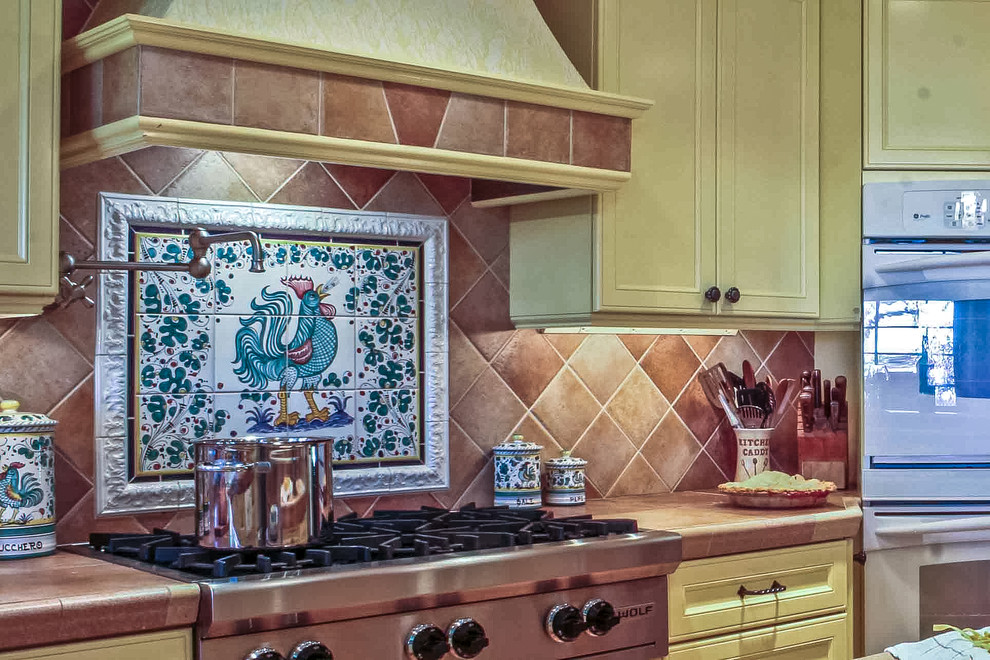 Photo of a large mediterranean l-shaped eat-in kitchen in Seattle with recessed-panel cabinets, yellow cabinets, tile benchtops, orange splashback, ceramic splashback, with island, white appliances and light hardwood floors.