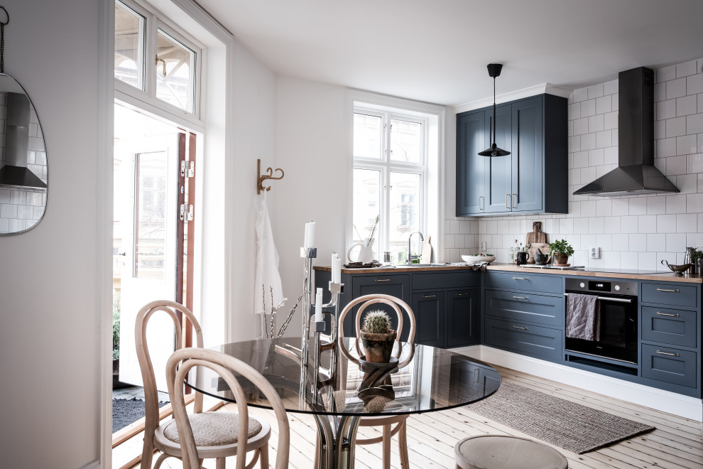 Photo of a scandinavian l-shaped eat-in kitchen in Gothenburg with shaker cabinets, blue cabinets, wood benchtops, white splashback, stainless steel appliances, light hardwood floors, no island, beige floor and brown benchtop.