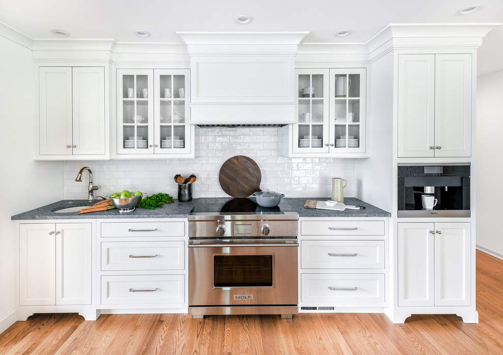Example of a large transitional l-shaped light wood floor eat-in kitchen design in Philadelphia with a farmhouse sink, flat-panel cabinets, white cabinets, soapstone countertops, white backsplash, ceramic backsplash, paneled appliances, an island and gray countertops