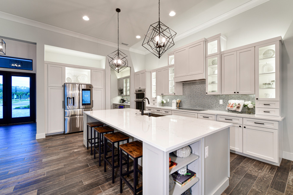 Mid-sized transitional l-shaped porcelain tile and brown floor eat-in kitchen photo with a farmhouse sink, shaker cabinets, white cabinets, quartz countertops, metallic backsplash, glass tile backsplash, stainless steel appliances, an island and white countertops