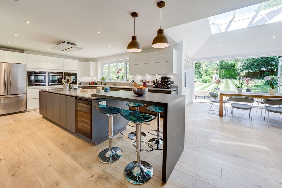 This is an example of a large contemporary eat-in kitchen in London with flat-panel cabinets, light hardwood floors, beige floor, white cabinets and stainless steel appliances.